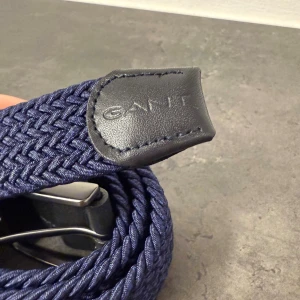 Marinblå flätat bälte från GANT - Snyggt marinblått flätat bälte från GANT med detaljer i svart skinn och en stilren metallspänne. Bältet har en modern och sportig look med tydlig struktur i materialet. Perfekt för att lyfta din outfit med en klassisk touch.