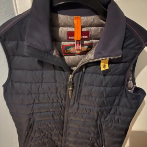 Mörkblå dunväst Parajumpers S - Snygg mörkblå dunväst från Parajumpers i storlek S. Västen har quiltad design, hög krage och dragkedja framtill. Insidan är ljusgrå och har en orange detalj vid nacken. Perfekt för lager-på-lager och chill stil.