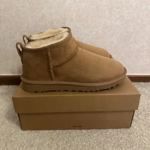 UGG Classic Mini bruna boots - Mysiga UGG Classic Mini boots i ljusbrun mocka med fluffigt fårskinnsfoder. Skorna har rund tå, platt sula och dragflik bak för enkel påtagning. Perfekta för kalla dagar och ger en chill vibe till din outfit.