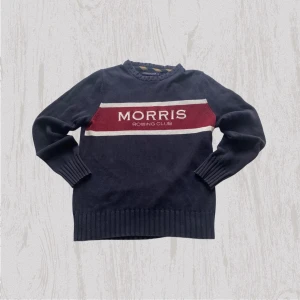 Morris Rowing Club stickad tröja - Mörkblå stickad tröja från Morris Rowing Club med bred vinröd och vit rand över bröstet och broderad logga. Långärmad med ribbade muddar och Union Jack-patch på ena ärmen. Perfekt för dig som gillar klassisk stil med sportig touch.