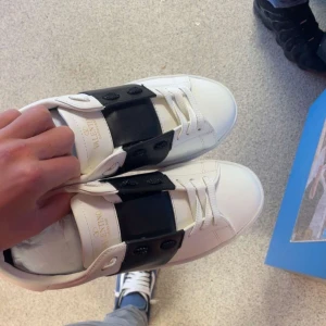 Valentino Garavani vita sneakers - Säljer ett par fräscha vita Valentino Garavani sneakers med bred svart läderdetalj över snörningen. Skorna har rund tå, platt sula och coola nitar på hälen. Perfekt för dig som gillar stilrena och lyxiga sneakers med unik design.