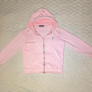 Ljusrosa hoodie från Polo Ralph Lauren - Snygg ljusrosa hoodie från Polo Ralph Lauren med dragkedja och huva. Tröjan har ett blått Polo-logo broderat på bröstet, två fickor fram och ribbade muddar. Perfekt för chill dagar och enkel att matcha med jeans eller leggings. Pris kan diskuteras