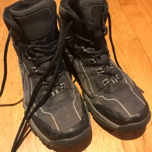 Svarta vattentäta kängor med snörning stl 40 - Säljer ett par svarta kängor med högt skaft och snörning. Skorna är vattentäta och har grå detaljer samt mönstrade partier på sidorna. Robust platt sula och rund tå, perfekta för höst och vinter. Materialet är syntet och textil.