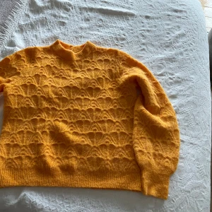 Orange stickad tröja från Object - Superfin gul stickad tröja från Object med vackert strukturmönster och två glittriga knappar i nacken. Tröjan har lång ärm, ribbade muddar och en liten öppning bak vid halsen. Perfekt för dig som vill ha något färgglatt och mysigt!