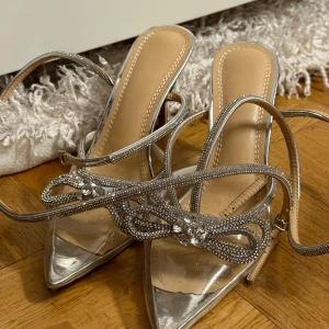 Silvriga klackpumps med strassdetaljer - Säljer ett par glittriga silvriga pumps med spetsig tå och hög klack. Skorna har tunna remmar dekorerade med strass och en snygg rosettdetalj framtill. Perfekta för dig som vill sticka ut med extra bling. Snygg design med öppen häl och spänne vid ankeln.
