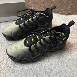 Nike Vapormax Plus svart/grå sneakers - Nike Vapormax Plus sneakers i svart och grått med neon-gula detaljer. Snygg futuristisk design med synlig Vapormax-sula och snörning. Ovandelen har en gradient från grått till svart och coola plastdetaljer längs sidorna. Perfekt för dig som gillar streetwear och unik stil.