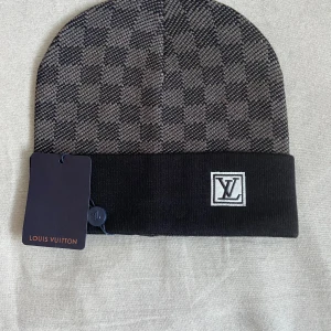 Rutig mössa från Louis Vuitton - Snygg mössa från Louis Vuitton i svart och grått med klassiskt rutigt mönster och vikt kant. LV-logga framtill på en patch. Tillverkad i mjukt material som håller dig varm och stilsäker under kalla dagar.