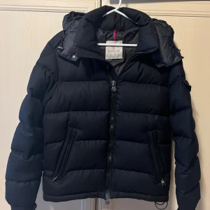 Svart Moncler Montgenevre Jacka - Säljer en svart pufferjacka från Moncler med avtagbar huva och två snedställda fickor med dragkedja. Jackan har klassisk Moncler-logga på dragkedjorna och är vadderad för extra värme. Perfekt för kalla dagar och riktigt snygg streetstil. Storlek 1, mycket bra skick.