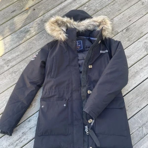 Svart vinterjacka Seaside - Svart vinterjacka från Seaside med huva och fuskpäls runt kanten. Jackan har stora fickor framtill, dragkedja och knappar. Insidan är fodrad för extra värme och huvan har mjukt teddyfoder. Använd fåtal gånger, inga hål eller smuts på jackan. 