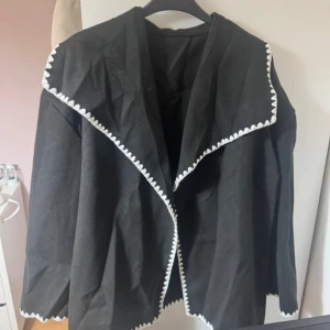 Svart kortkappa från SHEIN - Svart kappa från SHEIN i storlek L med bred krage och dekorativ vit kant längs krage och nederkant. Jackan är tillverkad i 100% polyester och har en loose passform med långa ärmar. Perfekt för dig som vill sticka ut med en unik detalj. Har använts 1 gång, säljs då den inte passade mig. 