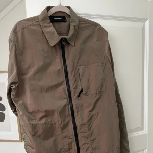 Grön overshirt Peak Performance - Snygg grön reflekterande overshirt från Peak Performance med rak passform och klassisk krage. Jackan har en bröstficka, hel dragkedja framtill och är tillverkad i lätt polyester. Perfekt för lager-på-lager och stilren look. Väldigt unik då den skiftar färg i olika ljus. Köpt för 2499. Använd 3-5 gånger och väl omhändertagen. 