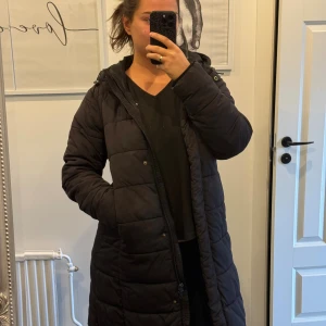 Svart lång pufferjacka med huva - Säljer en svart, lång pufferjacka med huva och dragkedja. Jackan har quiltad design, knappar framtill och två fickor på sidorna. Perfekt för kalla dagar och har en loose passform som gör den skön att bära över en hoodie eller tröja. Storlek M men passat även L/XL