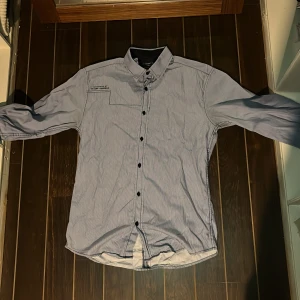 Randig skjorta från Selected Homme - Snygg skjorta från Selected Homme i ljusblått och vitt med tunna ränder. Klassisk krage, svarta knappar och lång ärm. Skjortan är i 100% bomull och har en diskret bröstdetalj med broderad text. Perfekt för en clean och stilren look.