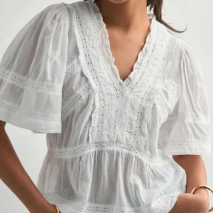 Vit blus med spets från Neo Noir - Superfin vit blus från Neo Noir med fina spetsdetaljer, v-ringning och volangkant. Blusen har korta, vida ärmar och är gjord i ett lätt och luftigt bomullstyg. Perfekt för en fräsch och somrig look!