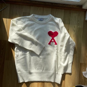 Vit stickad tröja från Ami Paris - Säljer en vit stickad tröja från Ami Paris med röd hjärt- och A-logga på bröstet. Tröjan har rund halsringning, långa ärmar och ribbade muddar. Perfekt för dig som gillar stilrena plagg med en twist.