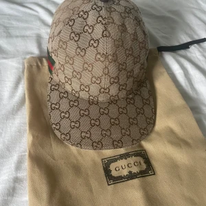 Gucci keps med GG-mönster och grön/röd detalj - Säljer en beige keps från Gucci med klassiskt GG-monogram över hela kepsen. Kepsen har en böjd skärm och en ikonisk grön/röd rand på sidan. Kommer med en dustbag. Använt några få gånger. Kontakta gärna om ni har frågor. Pris kan diskuteras.