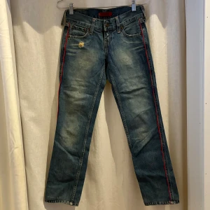 Levi's Patty Anne jeans med röd revär - Blå Levi's Patty Anne jeans med slim fit och raka ben. Snygg röd revär längs sidorna och slitning vid fickan framtill. Klassisk femficksmodell i jeansmaterial, perfekt för dig som gillar en cool och avslappnad stil. Som du ser på 4 bilden är en jeanshälla trasig men går att laga såklart.      Benlängd: 71cm