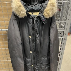 Svart parkas från Woolrich - Svart parkas från Woolrich med huva och beige päls. Jackan har dragkedja och knappar framtill, stora fickor och en varm, vadderad insida. Perfekt för kalla dagar och har en klassisk, stilren look. Storlek M