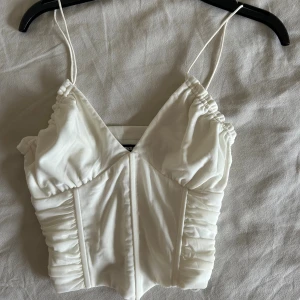 Vit korsett-topp från BikBok - Supersnygg vit korsett-topp från BikBok med tunna axelband och rynkade detaljer längs sidorna. Toppen har v-ringning och markerade sömmar framtill. Perfekt att styla med jeans eller kjol. Nypris: 399 kr