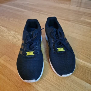 Svarta Adidas sneakers med Torsion - Snygga svarta Adidas sneakers med klassiska tre ränder på sidan och vit sula. Ovandelen är i mesh för extra andningsförmåga och skon har snörning samt en gul Torsion-detalj på plösen. Perfekta för en sportig och stilren look.