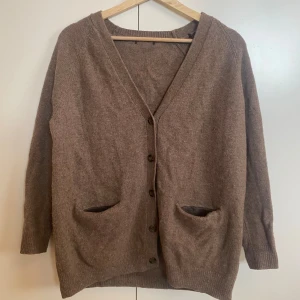 Brun stickad kofta med knappar - Mysig brun stickad kofta med v-ringning och knäppning framtill. Koftan har två praktiska fickor och långa ärmar. Enkel och tidlös design som passar till det mesta. Gjord i mjuk ull.