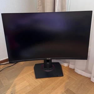 ASUS TUF Gaming VG27VQ - ASUS TUF Gaming VG27VQ är en 27-tums böjd gamingmonitor med robust design och stabil fot. Skärmen är tillverkad juni 2021 och har en modern, stilren look utan synliga defekter eller slitage. Perfekt för gaming med hög prestanda och snygg estetik.