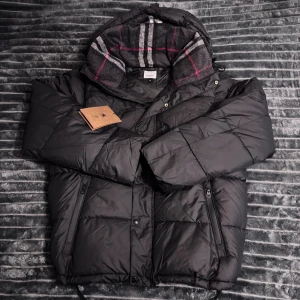 Burberry jacka - Säljer en svart pufferjacka från Burberry med avtagbara ärmar så den kan bäras som väst. Jackan har dragkedja, knappar och två fickor med dragkedja. Insidan av huvan har ett grått, vitt och rött rutigt mönster. Perfekt för kalla dagar och riktigt snygg streetstil.