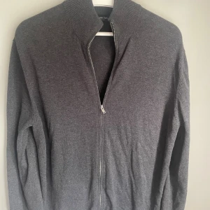 Grå kofta med dragkedja Massimo Dutti - Snygg mörkgrå kofta från Massimo Dutti med ribbad krage och hel dragkedja framtill. Tröjan har långärmad passform och är tillverkad i mjukt material som känns skönt mot huden. Perfekt för dig som gillar stilrena och enkla plagg.