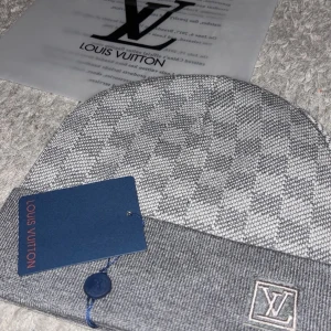 Louis Vuitton rutig mössa i grått - Snygg mössa från Louis Vuitton med klassiskt rutigt mönster i grå toner. Mössan har en uppvikt kant med LV-logga framtill och är tillverkad i mjukt stickat material. Finns i flera färger, men denna är grå och passar perfekt för att lyfta din streetstyle.
