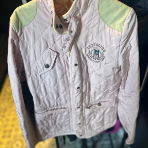 Quiltad rosa jacka från Lexington - Quiltad ljusrosa jacka från Lexington med broderad logga på bröstet, ljusbeiga axelpartier och detaljerade knappar. Jackan har hög krage, tryckknappar och fickor framtill. Perfekt för dig som gillar klassisk och sportig stil.