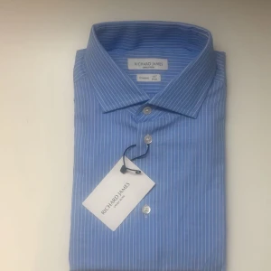 Richard James Savile Row – Classic skjorta (helt ny) - Elegant ljusblå skjorta med vita ränder från Richard James Savile Row, känd för sin brittiska skräddarkvalitet.                                                 Modell: Classic fit.                                             Storlek: 15½” / 39 cm (S)                               Skjortan är helt ny, aldrig använd, och har originaltaggen kvar, även 100% bomull Nypris: cirka 2 600 kr Pris: 700 kr  En stilren och välsydd skjorta i klassisk passform, perfekt för både vardag och formella tillfällen.