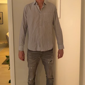 Randig skjorta från Euro Fashion - Klassisk skjorta från Euro Fashion med smala blå och vita ränder. Skjortan har lång ärm, bröstficka och knappar framtill. Tillverkad i bomull för en skön känsla och snygg passform. Perfekt för dig som gillar stilrena och tidlösa plagg.