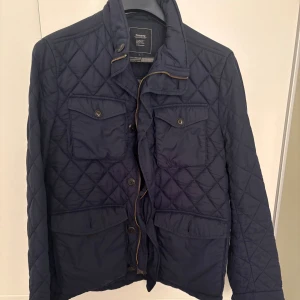 Mörkblå quiltad jacka från Boomerang - Snygg mörkblå quiltad jacka från Boomerang med klassisk krage, fyra fickor framtill och både dragkedja och knappar. Jackan har ett rutigt mönster och är tillverkad i polyester, perfekt för höst och vår.