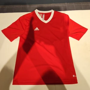 Röd Adidas fotbollströja L - Snygg röd fotbollströja från Adidas med vit V-ringad krage och Adidas-logga broderad på bröstet. Tröjan är kortärmad och tillverkad i lätt, funktionellt material som andas. Perfekt för träning eller match.