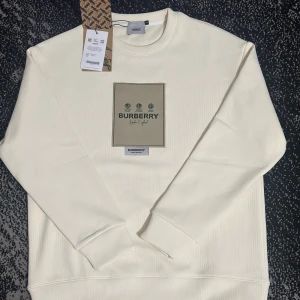 Vit sweatshirt från Burberry - Säljer en stilren vit sweatshirt från Burberry med klassisk logga framtill. Tröjan har rund halsringning, långa ärmar och ribbade muddar. Tillverkad i mjuk bomull för en skön känsla och clean look. Perfekt för dig som gillar exklusiva streetwear-vibbar.