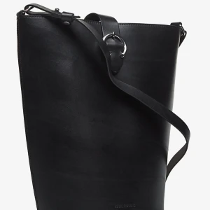 Svart axelväska i skinn från Royal Republiq - Stilren svart axelväska från Royal Republiq i slätt skinn. Väskan har en minimalistisk design med justerbar axelrem och silverfärgad metallring som detalj. Perfekt för dig som gillar en clean look och vill ha plats för allt du behöver. Nypris 2200 Aldrig använd