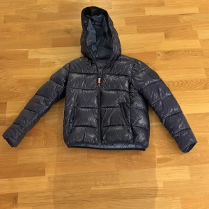 Mörkblå Vinterjacka från Save The Duck - Säljer en mörkblå Save The Duck pufferjacka med huva och dragkedja. Jackan har två snedställda fickor med dragkedja och quiltad design. Perfekt för kalla dagar och riktigt skön att bära. Passformen är normal och jackan är tillverkad i syntetmaterial. Storlek 12 år cirka 152-158 cm pris 599 kr