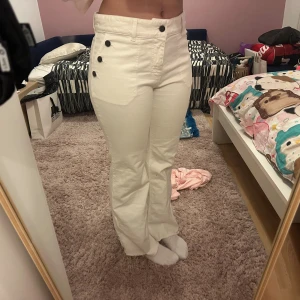 Vita bootcut byxor med knappar - Säljer ett par vita bootcut-byxor med hög midja och snygga svarta knappar längs sidan. Byxorna har råa kanter nertill och är tillverkade i ett mjukt bomullstyg. Perfekta för dig som vill sticka ut med en cool detalj i vardagslooken.