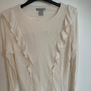 Vit långärmad topp med volang - Säljer en söt vit långärmad topp från H&M med volangdetaljer längs framsidan. Toppen har rund halsringning och är gjord i mjuk bomull, perfekt för en clean och stilren look.