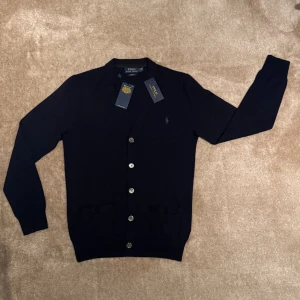 Marinblå kofta från Polo Ralph Lauren - Snygg marinblå kofta från Polo Ralph Lauren i slim fit med mjuk merinoull. Tröjan har v-ringning, långa ärmar, sex knappar framtill och två fickor. Diskret logga broderad på bröstet. Perfekt för dig som gillar klassisk och stilren look.