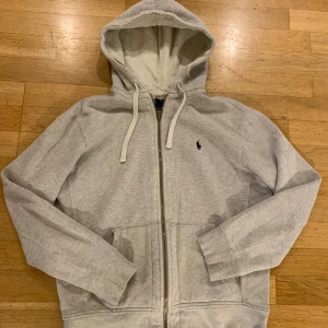 Grå hoodie från Polo Ralph Lauren - Snygg grå hoodie från Polo Ralph Lauren med klassisk logga på bröstet. Tröjan har huva med snörning, dragkedja framtill och två fickor. Tillverkad i mjuk bomull och har långa ärmar. Perfekt för chill dagar och enkel att matcha med jeans eller joggers. Några små hål och revor