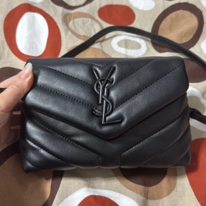 Svart quiltad axelväska från YSL - Snygg svart axelväska från Saint Laurent med ikoniska YSL-loggan framtill. Väskan är quiltad i skinn och har ett magnetlås samt innerfack med dragkedja. Perfekt storlek för det viktigaste och har en stilren, modern look.