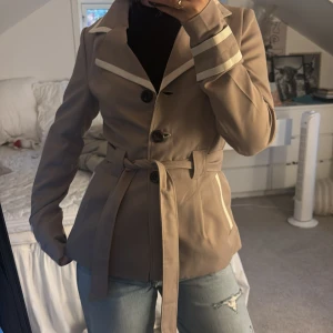 Beige trenchcoat med bälte och vita detaljer - Snygg beige trenchcoat med markerad midja och bälte. Jackan har vita detaljer längs krage, ärmslut och fickor samt stora knappar framtill. Klassisk krage och långärmad modell, perfekt för dig som vill ha en stilren look.