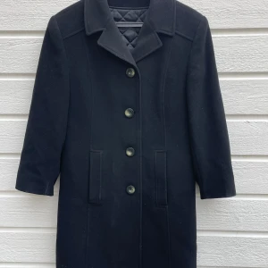 Svart kappa från Pabia i ull och kashmir - Stilren svart kappa från Pabia i en lyxig mix av ull och kashmir. Klassisk rak modell med krage, knäppning framtill med fyra knappar och två diskreta fickor. Ärmarna är av trekvartsmodell. Liten i storlek. Mer lik storlek S