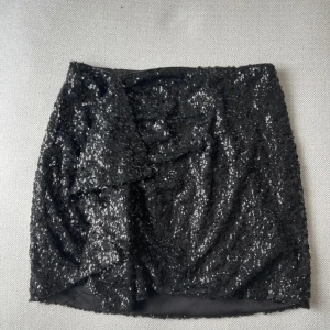 Svart paljettkjol med omlott - Supersnygg svart kortkjol täckt av glittrande paljetter och med omlott-detalj framtill. Kjolen har en figurnära passform. Perfekt för dig som vill sticka ut med en glammig look.