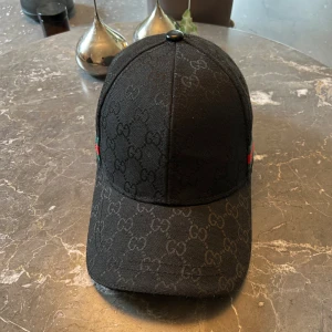 Svart keps med GG-mönster - Snygg svart keps med gg mönster. Justerbar rem baktill. Tillverkad i bomull med detaljer i polyester och elastan. 