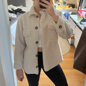 Beige teddyjacka med stora knappar - Säljer en beige teddyjacka. Jackan har stora bruna knappar framtill, två rejäla bröstfickor och mjukt, fluffigt material. Ganska tunn så kan användas både som jacka eller som overshirt/skjorta. Storlek 32 men passar en 342xs/s 