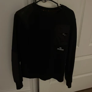 Svart fleece tröja Parajumpers YL - Svart långärmad fleece tröja från Parajumpers med cool patch på ärmen och bröstficka med dragkedja. Snygga detaljer med orange sömmar vid halsen och logga på bröstet. Perfekt för dig som gillar streetwear och vill ha något bekvämt och stilrent.