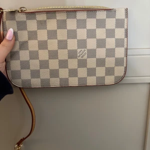  kuvertväska - Snygg kuvertväska i klassiskt Damier Azur-mönster. Väskan är beige och vit med rutigt mönster, har guldfärgad dragkedja och avtagbar läderrem. Perfekt storlek för det viktigaste och en ikonisk accessoar till din outfit.