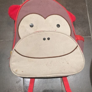 Ryggsäck med apa från Skip Hop - Söt ryggsäck från Skip Hop med apansikte framtill, röda detaljer och gula dragkedjor. Väskan har meshficka på sidan, justerbara axelband och gott om plats för böcker eller snacks. Perfekt för skolan eller utflykten!